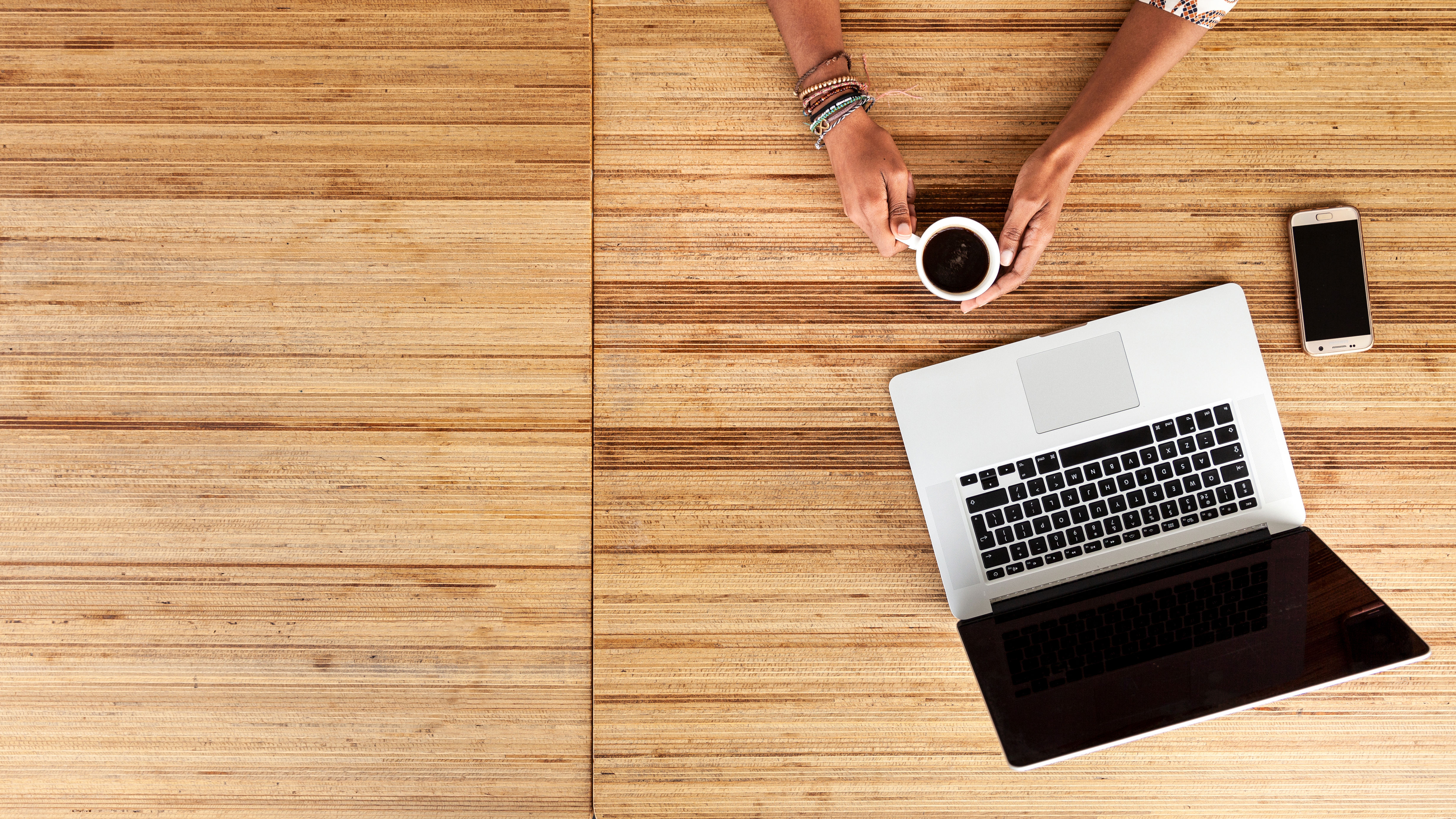 Een laptop staat op een houten tafelblad. Twee handen houden een kop koffie vast.