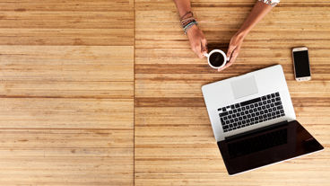 Een laptop staat op een houten tafelblad. Twee handen houden een kop koffie vast.