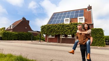 Iemand zit op de rug van een man. Samen kijken ze in de camera. In de achtergrond staat een woning met zonnepanelen op het dak.