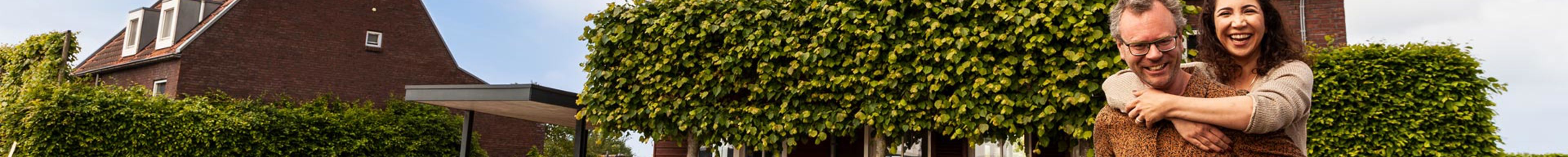 Koppel voor huis met zonnepanelen Iemand zit op de rug van een man. Samen kijken ze in de camera. In de achtergrond staat een woning met zonnepanelen die geplaatst zijn door Greenchoice.